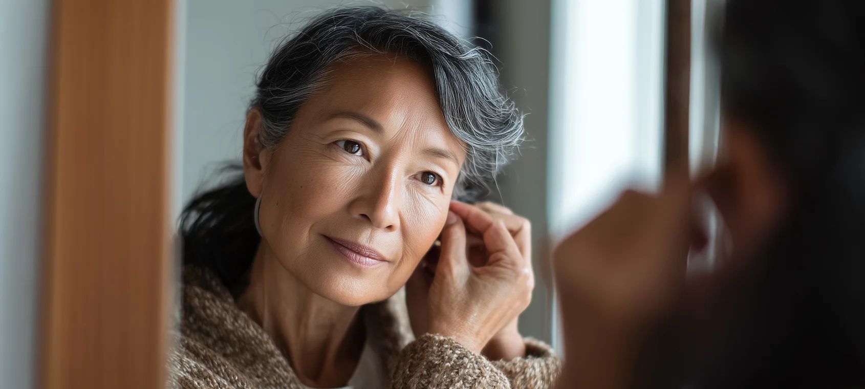 Elegant senior woman putting on earrings in front of mirror